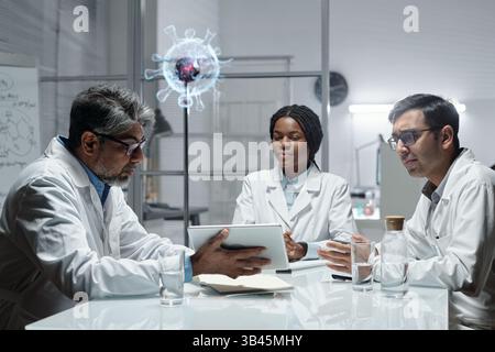 Group of scientists in lab coats collaborating around a table, sharing ideas and data on a digital tablet with holographic virus model in background Stock Photo