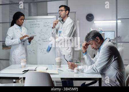 Group of scientists in lab coats sharing ideas and reviewing notes while standing near a whiteboard and table filled with research materials, coffee cups, and equipment Stock Photo