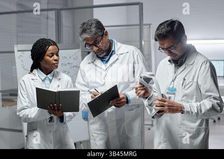 Group of three scientists wearing lab coats discussing research findings in laboratory environment, using various scientific instruments Stock Photo