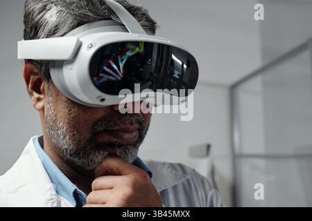 Middle-aged man in laboratory environment examining virtual content displayed in advanced VR headset thinking deeply wearing white lab coat. Modern technology in scientific research Stock Photo