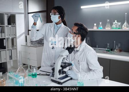 Scientist examining blue liquid in test tube while colleague observing through microscope in modern laboratory setting involving scientific research and analysis Stock Photo