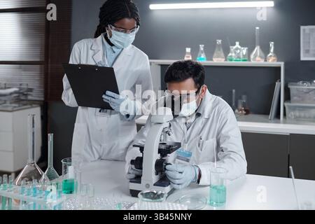 Scientists conducting research in modern laboratory with various glassware and equipment. One scientist is examining samples under microscope while another is taking notes on clipboard Stock Photo
