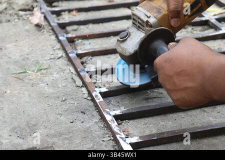 A worker is smoothing welded metal frame outdoors using a handheld angle grinder. Sparks are visible during metal work. Close-up of tool and hands. Stock Photo