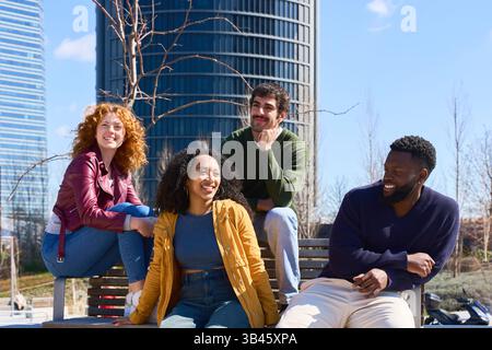 Four cheerful young multiracial businesspeople sitting on bench having fun talking and laughing during work break outdoors Stock Photo