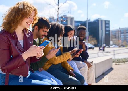 Group of diverse young students sitting in a row outdoors, engaging with smartphones while enjoying a sunny day in the urban landscape. People and tec Stock Photo