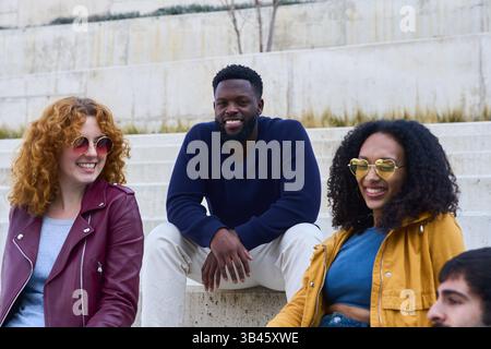 Multi ethnic group of students sitting together on concrete steps, enjoying their free time between classes Stock Photo