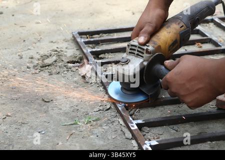 Close-up shot shows sparks flying as a dark-skinned person uses an angle grinder on a metal frame outdoors, focusing on the grinding process. Stock Photo