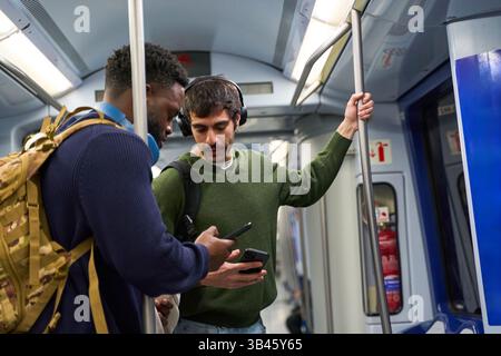 Two male students standing inside a moving subway train, focused on their smartphones while enjoying their commute through the urban landscape Stock Photo