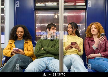 Four young students sitting together in a subway train, engaged with their smartphones while enjoying the urban commute and connecting online Stock Photo