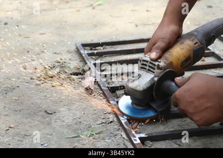 Close-up of dark-skinned metalworker using an angle grinder on steel frame, generating sparks in an outdoor setting. Shows hand and tool details. Stock Photo