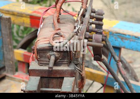 Focused shot displays an old rusty motor assembly with connecting bolts and wires on a weathered frame painted in sections of blue, yellow, and red. Stock Photo