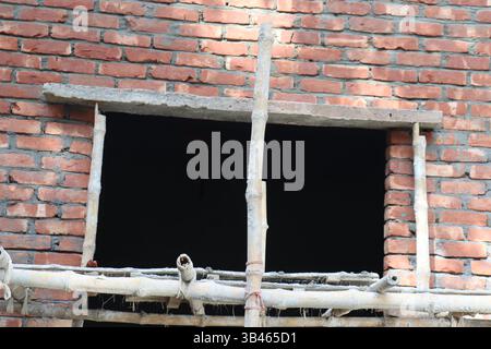 Unfinished brickwork shows a rectangular opening framed by bamboo scaffolding against an interior. Construction shows red bricks, gray mortar, bamboo, Stock Photo