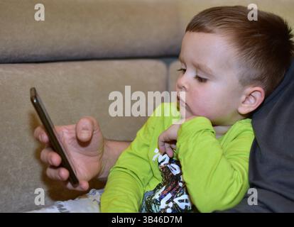 Toddler boy is watching video through smart phone with his dad. Child is looking at phone screen at home. Dad and son uses smart technologies at home. Stock Photo