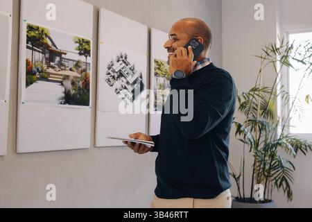 A dedicated project manager talks on the phone while examining design plans pinned on a wall. The setting portrays a professional environment with a f Stock Photo