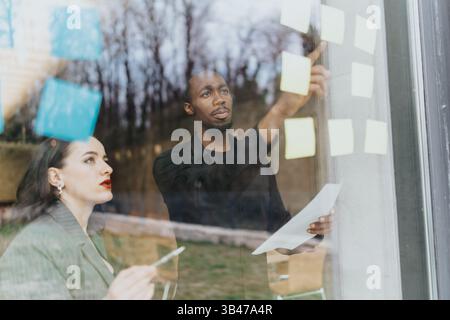 Two business colleagues collaborating on a project with sticky notes on a glass wall Stock Photo