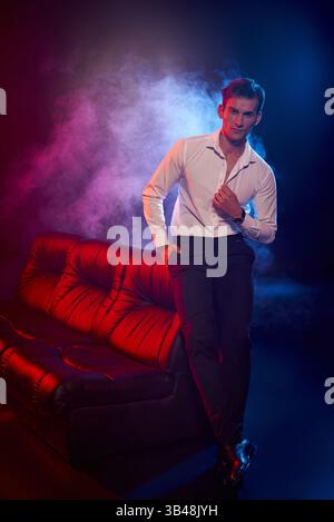Charismatic young man poses confidently in a stylish outfit, surrounded by vibrant smoke and light. Stock Photo