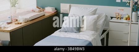 Hospital room featuring a bed with clean white linens and light blue blanket, accompanied by bedside table, window with blinds ushering sunlight and variety of medical equipment Stock Photo
