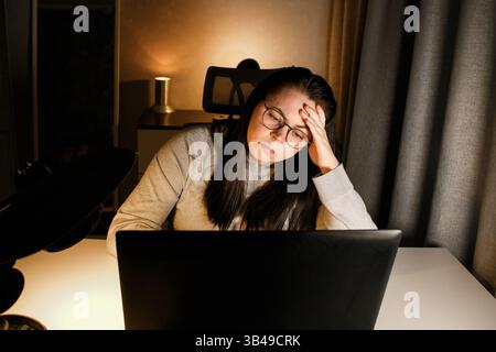 Woman working on computer late evening at home office. Concentrated female freelancer trying to solve business problems. Working and suffering from we Stock Photo