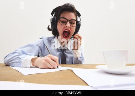 Frustrated young woman in office wear yelling into headset while taking notes with pen at messy desk against white background. Stock Photo
