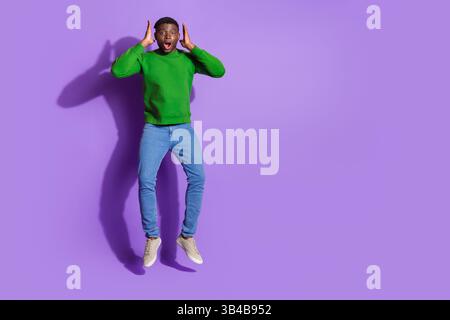 Joyful young man in casual green sweatshirt jumping with surprised expression against a vibrant purple background Stock Photo