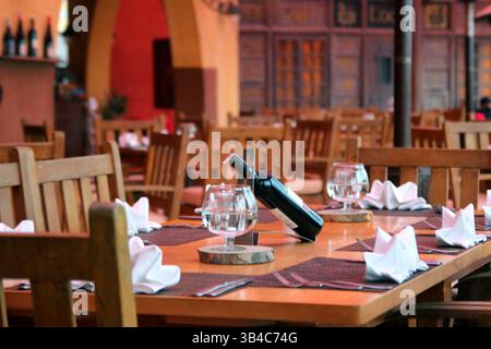 Stylish restaurant interior featuring a set table with wine bottle, water glasses, folded napkins, and wooden chairs in warm ambient lighting. Stock Photo