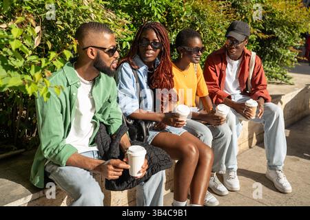 Lively group of African American college friends enjoying conversation with takeaway drinks in park Stock Photo