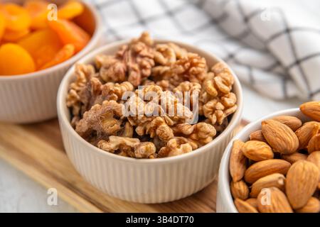 Three white ceramic bowls filled with almonds, walnuts, and dried apricots, arranged on a wooden cutting board, offer a healthy and delicious snack or Stock Photo