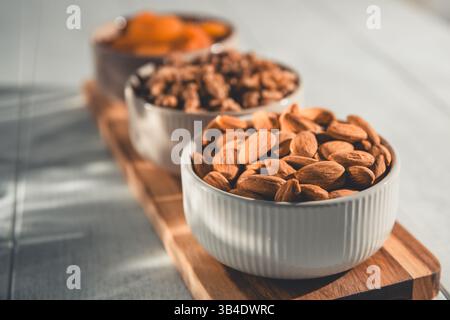 Three white ceramic bowls filled with almonds, walnuts, and dried apricots, arranged on a wooden cutting board, offer a healthy and delicious snack or Stock Photo