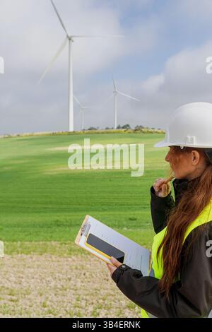 Female engineer inspecting wind turbines in green field, analyzing data for sustainable energy Stock Photo