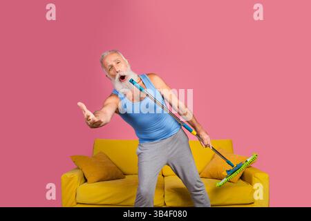 Senior man singing passionately using a mop as a microphone in a colorful living room Stock Photo