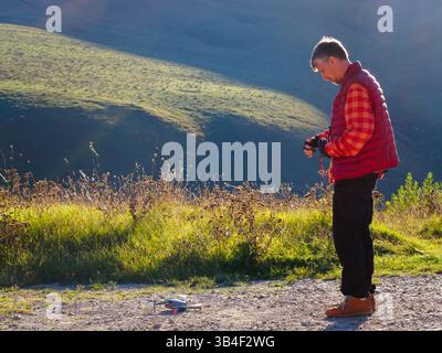 Man in red plaid vest operates drone on mountainside Stock Photo