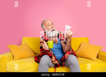 Elderly man sitting on a yellow sofa with a pink background, holding a cup and tissue, wrapped in a plaid blanket, appearing thoughtful Stock Photo