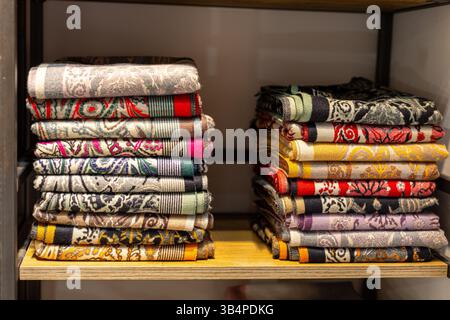 Stacks of beautifully folded fabrics with detailed patterns are neatly arranged on wooden shelves in a textile shop. The colorful variety highlights c Stock Photo