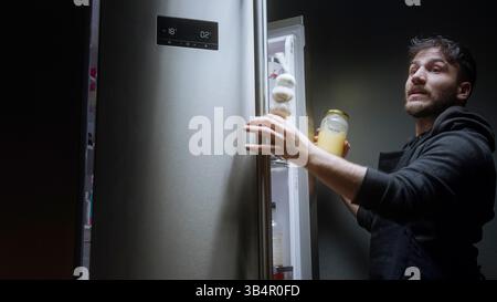 Young Man Holding a Jar and Looking Inside Refrigerator Late at Night in Dim Kitchen Stock Photo