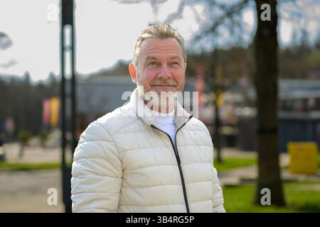 An older man with a friendly expression stands outdoors in a sunny park, looking slightly upwards with a thoughtful smile. Stock Photo