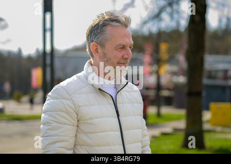 A thoughtful older man stands in a sunlit park wearing a light beige puffer jacket. Stock Photo