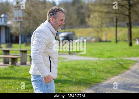 A casually dressed older man walks through a sunny park, smiling warmly. Stock Photo