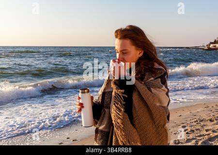 Young woman standing by the ocean shore, wrapped in cozy blankets, enjoying a hot drink from a thermos on a breezy day at the beach Stock Photo