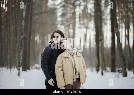 Couple in a snowy forest with the woman leaning on the man, both in winter attire among towering trees Stock Photo