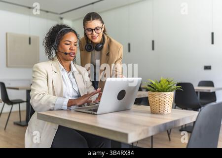Two female telemarketers wearing headsets are collaborating on a laptop in a brightly lit modern office, showcasing teamwork and communication Stock Photo