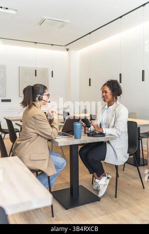 Two women recording a podcast episode, using professional microphone and headphones, sitting at a table in a modern studio Stock Photo