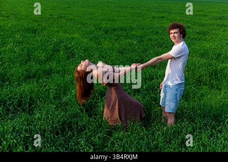 Joyful young couple holding hands and laughing together in a lush green meadow, the woman leaning back playfully while the man smiles warmly, enjoying Stock Photo