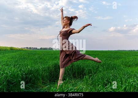 Carefree young woman in a floral dress leaping gracefully in a lush green meadow with a bright sky and soft clouds in the background Stock Photo