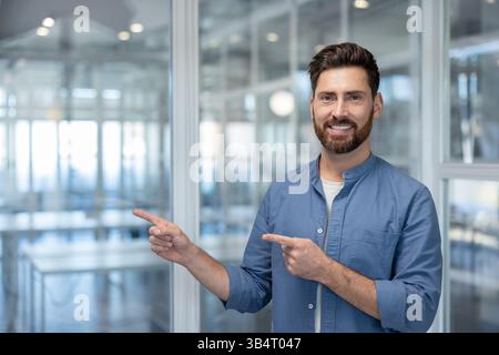 A smiling, bearded man points with both hands, suggesting something is to his side in an office setting. Stock Photo