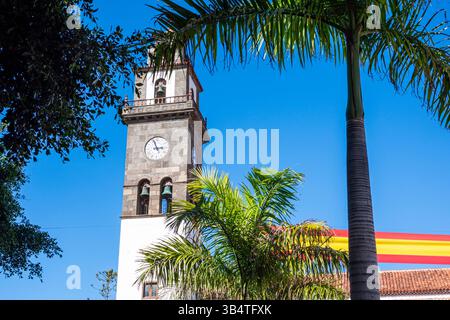 A large clock tower with a cross on top stands in front of a red and yellow banner. The banner is attached to a pole and is hanging over the building. Stock Photo