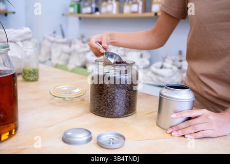 Saleswoman scooping roasted coffee beans from glass jar into metal can in bulk food store Stock Photo