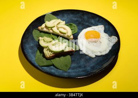 Breakfast dish of scrambled eggs and sandwiches with avocado and spinach on a blue plate. Yellow background minimalism. Stock Photo