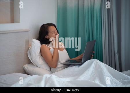 tired woman working on a laptop in the evening in bed. yawns, covers his mouth with his hand. Stock Photo