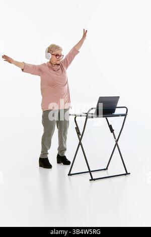 Senior woman dancing in headphones with arms raised, repeat moves during online lessons against white background. Active aging. Stock Photo