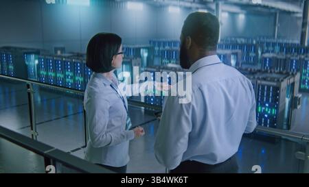 Big Data Center Multiethnic Specialists with Digital Tablet Talking, Shaking Hands, Looking at Camera. Server Racks for Cloud Computing Technology. Concept of Information Digitalization, Web Service. Stock Photo